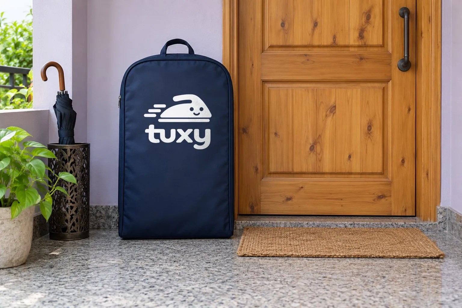 A navy Tuxy garment bag beside a natural pine door with an iron handle on a soft lilac wall, with an umbrella stand with one folded umbrella and a jute doormat.